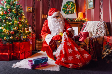 Portrait of Santa Claus - with a white beard, Santa's hat, with gifts near the Christmas tree and fireplace on an armchair on Christmas Eve and New Year.