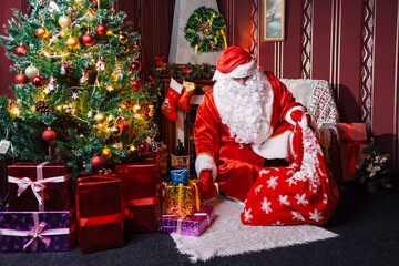 Portrait of Santa Claus - with a white beard, Santa's hat, with gifts near the Christmas tree and fireplace on an armchair on Christmas Eve and New Year.