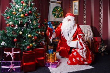 Portrait of Santa Claus - with a white beard, Santa's hat, with gifts near the Christmas tree and fireplace on an armchair on Christmas Eve and New Year.
