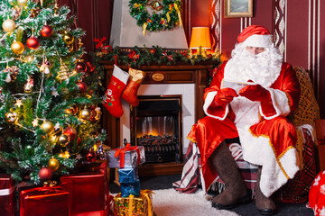 Portrait of Santa Claus - with a white beard, Santa's hat, with gifts near the Christmas tree and fireplace on an armchair on Christmas Eve and New Year.