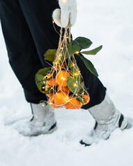 The string bag contains tangerines with leaves and a garland. A girl in mittens carries tangerines home. Preparing for Christmas dinner. Meeting the new year. Waiting for a miracle. Christmas food.