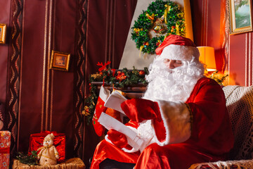 Portrait of Santa Claus - with a white beard, Santa's hat, with gifts near the Christmas tree and fireplace on an armchair on Christmas Eve and New Year.