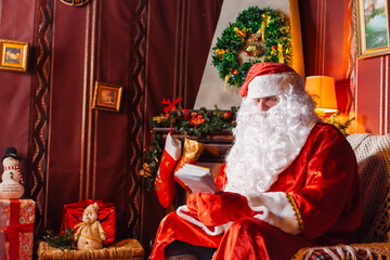 Portrait of Santa Claus - with a white beard, Santa's hat, with gifts near the Christmas tree and fireplace on an armchair on Christmas Eve and New Year.