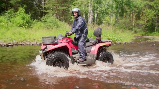 Slowmo tracking of bearded man in helmet driving red four-wheeler through river in forest