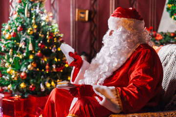 Portrait of Santa Claus - with a white beard, Santa's hat, with gifts near the Christmas tree and fireplace on an armchair on Christmas Eve and New Year.