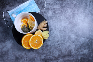 White cup of tea with lemon, ginger, orange., jam and medical mask on the dark grey background. Healthcare. Vitamin cocktail.