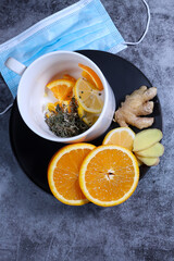 White cup of tea with lemon, ginger, orange., jam and medical mask on the dark grey background. Healthcare. Vitamin cocktail.