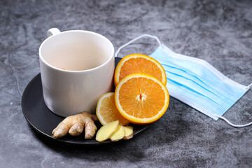 White cup of tea with lemon, ginger, orange., jam and medical mask on the dark grey background. Healthcare. Vitamin cocktail.