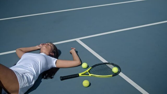 Tired depressed young female tennis player lies on outdoor tennis court with a lot of balls after hard training at sunset. Top view. Motivational video, power and result
