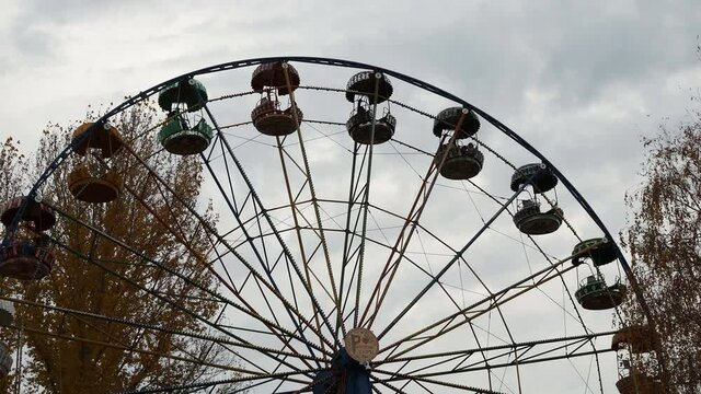 Cloudy autumn day. Sky with dark clouds. Bottom view. Ferris wheel in gray silver metal in amusement park. Ferris wheel is slowly moving in circle.