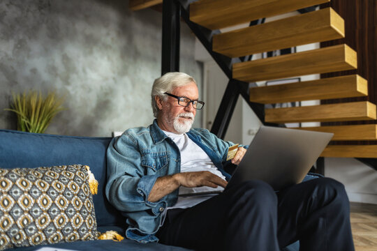 Modern Elderly Person Place An Order For Purchase Of Goods Online Sitting On The Couch At Home. Happy Senior Male Smiling And Looking At The Laptop Shopping Online With Credit Card