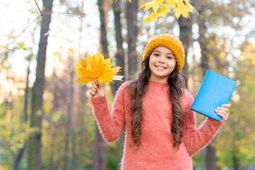 Happy small child hold school book collecting fall leaves in autumn park outdoors, library
