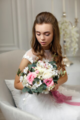 beautiful tender young bride with wedding makeup and long curly hair holding flower bouquet and sitting on armchair in white studio interior