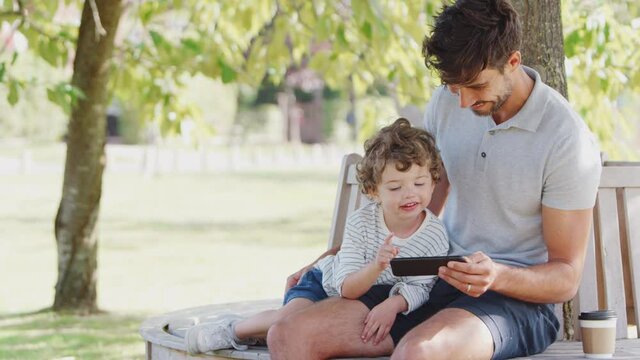 Father sitting on park bench under tree with son looking at video game on mobile phone together - shot in slow motion
