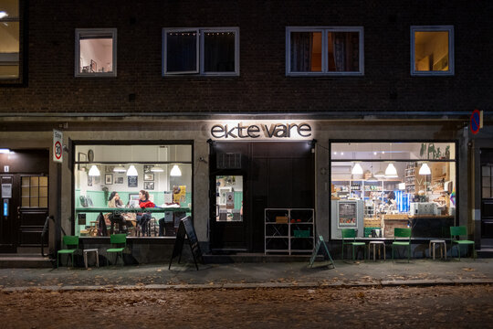 Oslo, Norway - 11/03/2020: Ekte Vare - Organic Groceries -  Storefront At Night With Interior Lights And View From The Street At Sagene With Green Chairs Outdoors And People Sitting By The Window