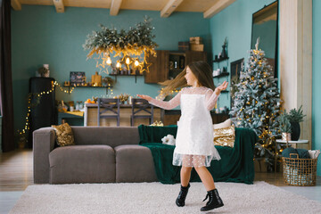 A cheerful little girl in a white dress is spinning in front of a Christmas tree in a cozy room