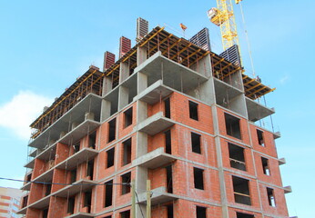 A multi-storey apartment building is being built with brick and concrete. Construction of the building against the blue sky. The tower crane is yellow