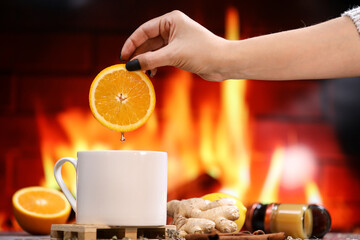 A woman's hand puts a slice of orange into white tea cup with lemon, ginger, orange and jam against the background of a burning fireplace. Healthcare. Vitamin cocktail.	