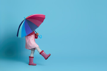 happy little child girl with multicolored umbrella in pink rain coat and rubber boots on blue...