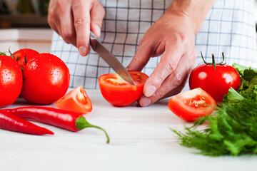 Male hand with a knife cuts a fresh ripe tomato on wooden white board. Process of making vegetable salad . Healthy food concept. Close-up.