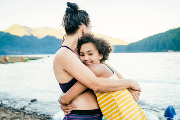 Fototapeta na wymiar Smiling teen girl hugging mom while she looks away at lake in forest at sunset