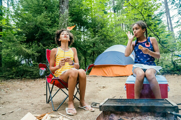 Teen girl blowing fire off of toasted marshmallows while sitting in campground by campfire, while sister licks her finger