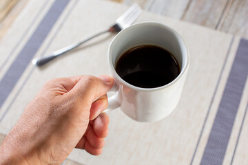 A view looking down onto a hand holding a mug of coffee, over a plate setting, in a restaurant or kitchen setting.