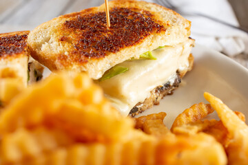 A closeup view of a chicken melt sandwich, with crinkle cut french fries in the foreground, in a restaurant or kitchen setting.