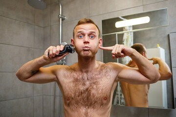 A man shaves his face with an electric razor in front of a mirror. Skin irritation. Bath procedure