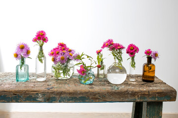 Autumn asters in small pharmacy bottles instead of vases on a long wooden bench against white wall.