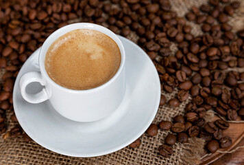 White cup of frothy coffee and sprinkled beans on sacking. Close-up. Top view.