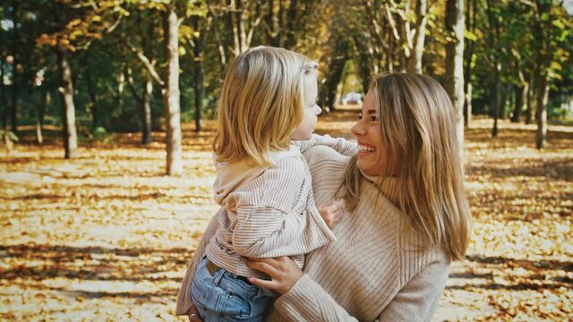 Young mom is smiling, holding little daughter in arms and tickling her during their walk in autumn park