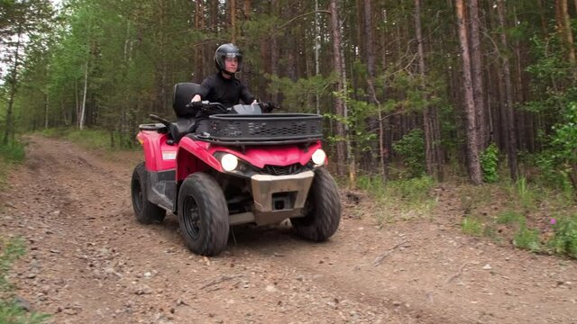 Handheld shot of man in helmet driving red quad bike downhill along dirt road in forest