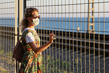 Young woman wearing protective mask looks through the grille at the sun and sea. Concept of the desire for freedom during pandemic