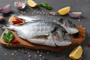Fresh ready to cook raw fish dorado with ingredients and seasonings like basil, lemon, salt, pepper, red onion and garlic on wooden board on dark background.