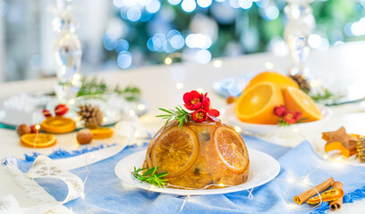 Traditional English Christmas pudding decorated with oranges and flowers in a festive table setting with fruits, spices, and lights of Christmas tree on background. Copy space. Magical mood. Wide card