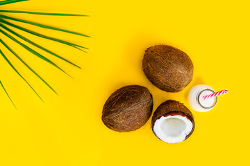 Obraz na płótnie Canvas Top view coconut water drink, milk in a bottle with straw, and fresh coconuts with a green palm leaf on the yellow background. Summer exotic refreshment. Natural plant based food. Flatlay. Copy space.