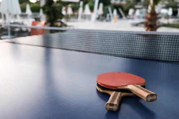 Closeup view of two table tennis rackets with net on blue surface. Selective focus, hotel or park in blurred background. Fun competition for two or entertainment on vacation. Active lifestyle concept