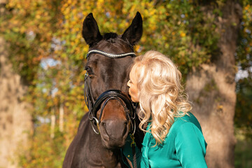 Obraz premium Young blond woman in green dress, kissing a brown horse. In the forest at autumn in golden color. love horses