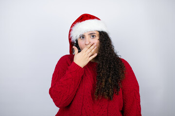 Young beautiful woman wearing a Santa hat over white background surprised covering the mouth