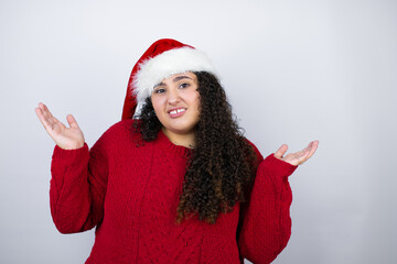 Young beautiful woman wearing a Santa hat over white background clueless and confused expression...