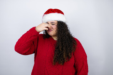 Young beautiful woman wearing a Santa hat over white background smelling something stinky and disgusting, intolerable smell, holding breath with fingers on nose