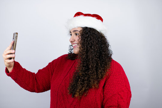 Young beautiful woman wearing a Santa hat over white background taking a selfie with her phone