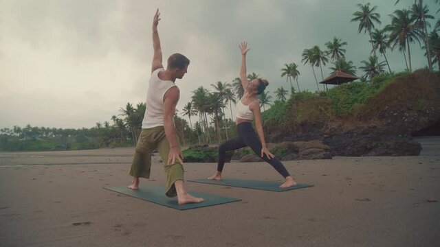 Full over shoulder of male sun-tanned muscular yoga teacher showing asana to female apprentice standing on mats. People exercising on uninhabited sandy beach by rocky hill with palm trees on it