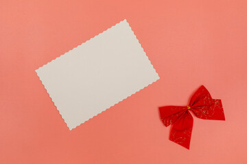 Blank white postcard and red bow decoration on pink background. Christmas, New Year, Valentine's Day, International Women's Day or Mother's Day composition. Flat lay with copy space.