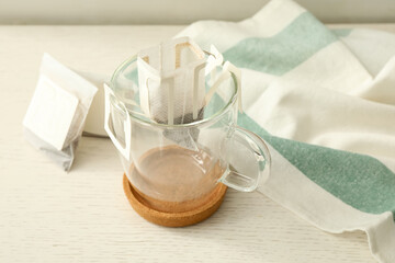 Drip coffee bags and glass cup on white wooden table, closeup