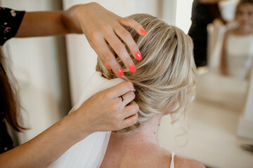 Fototapeta premium A professional hairdresser makes a wedding hairstyle to a young beautiful blonde woman on the wedding day. Morning preparations for the bride to meet with the groom. Ideal bride concept
