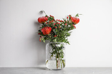 Bouquet with beautiful red protea flowers in glass vase on grey table against white background