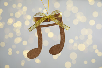 Wooden music note with golden bow hanging on light grey background with blurred Christmas lights
