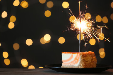 Cake with burning sparkler on black table against blurred festive lights. Space for text